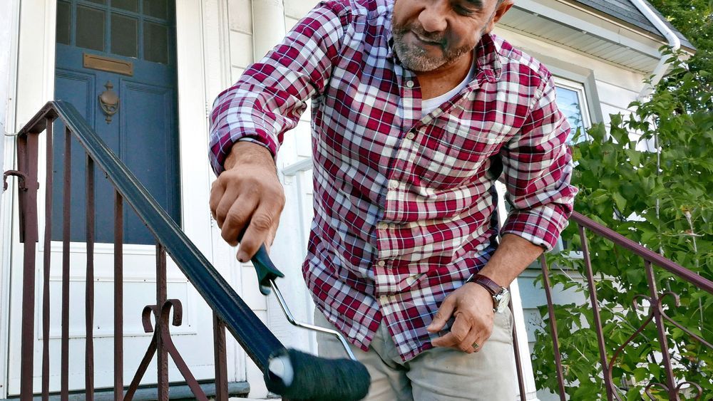 Mauro Henrique using a roller to paint the top of a rusty hand rail.